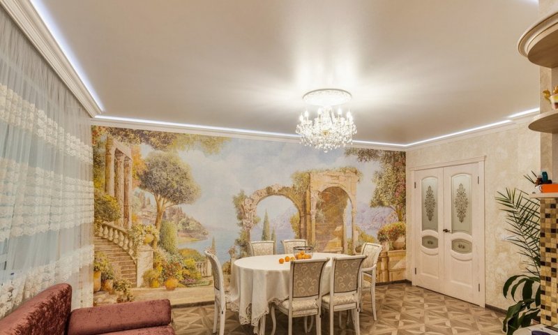 Матова натяжна стеля у вітальні будинку з парящим ефектом і LED-підсвіткою додає простору легкості та елегантності. Приховане освітлення по периметру створює м'яке, розсіяне світло, підкреслюючи класичний стиль інтер’єру. Центральна кришталева люстра надає розкішного акценту, ефектно освітлюючи вишукану меблеву групу та декоративне панно з середземноморським пейзажем. Легкі ажурні штори, благородна палітра кремових і золотистих відтінків, різьблені меблі та класичний паркет створюють атмосферу витонченого стилю та затишку, роблячи простір візуально більшим і гармонійним.