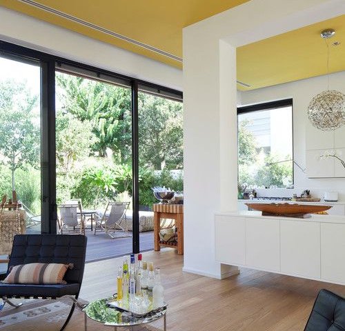 Натяжна стеля у вітальні у теплому жовтому відтінку додає інтер'єру світла та затишку. Завдяки великим панорамним вікнам простір наповнюється природним освітленням, а поєднання світлих стін і дерев'яної підлоги створює атмосферу природної гармонії.