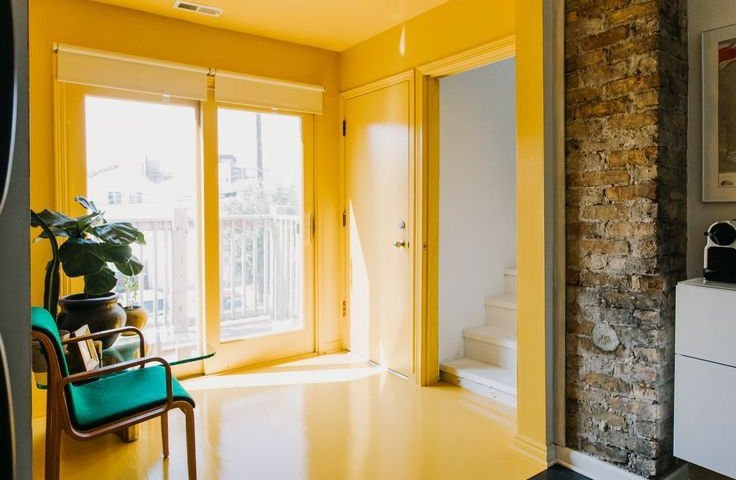 Глянцева натяжна стеля у передпокої яскравого жовтого кольору візуально розширює простір і додає сонячної енергії інтер’єру. Поєднання з натуральними матеріалами, такими як цегляна стіна та дерев’яна підлога, створює стильний сучасний вигляд.